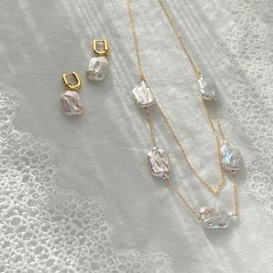 May include: A gold necklace and matching earrings set featuring irregular, white baroque pearls. The necklace has a double strand design. The earrings are small hoops. The jewelry is displayed on a white, textured surface.