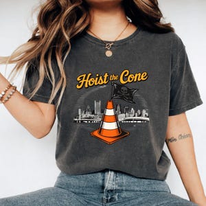 May include: Dark gray t-shirt with the text "Hoist the Cone" in yellow and a graphic of a traffic cone with a skull and crossbones flag. The design includes a city skyline.