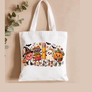 May include: A white canvas tote bag featuring a colorful graphic print of a smiling apple wearing a witch hat, a stack of books, a pencil, a ghost with a bow, and a pumpkin, all surrounded by small autumn leaves, stars, and bats.