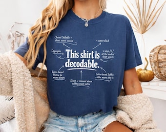 May include: A navy blue short-sleeve t-shirt featuring white text that reads "This shirt is decodable." with arrows pointing to linguistic terms including "Closed Syllable," "r-controlled," "Digraphs," "s says /z/," "Latin-based Prefix de-," and "Latin-based Suffix -able."