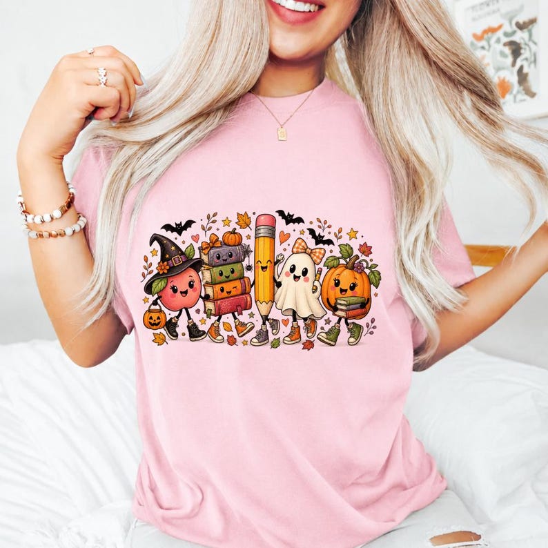 May include: A light pink t-shirt featuring a colorful graphic print of a smiling apple wearing a witch hat, a stack of books, a pencil, a ghost with a bow, and a pumpkin, all surrounded by small autumn leaves, stars, and black bats.