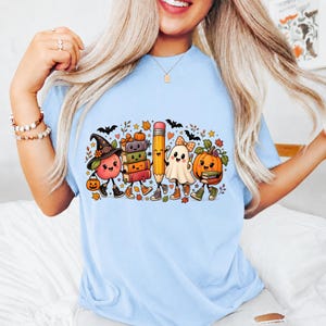 May include: A light blue t-shirt featuring a colorful graphic print of a smiling apple wearing a witch hat, a stack of books, a pencil, a ghost, and a pumpkin. The design includes small bats, autumn leaves, and stars scattered around the characters.