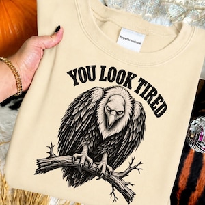 Può includere: Una felpa girocollo beige con un'illustrazione grafica nera di un avvoltoio appollaiato su un ramo. Sopra l'uccello, il testo "YOU LOOK TIRED" è scritto in un carattere grassetto e arcuato. L'etichetta interna bianca sul collo riporta la scritta "hypethreadmok".