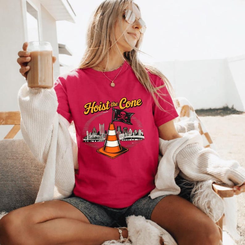 May include: A magenta t-shirt with the text "Hoist the Cone" in yellow script. The graphic features a traffic cone with a skull and crossbones flag, and a city skyline. The shirt is worn by a person.