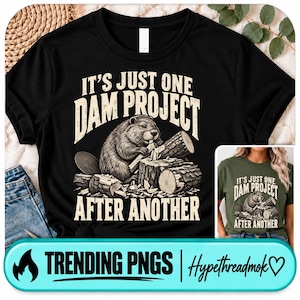 May include: A black short-sleeved t-shirt featuring a graphic of a beaver chewing on a log with the text IT'S JUST ONE DAM PROJECT AFTER ANOTHER in a beige font. The design includes wood logs and wood chips surrounding the beaver illustration.