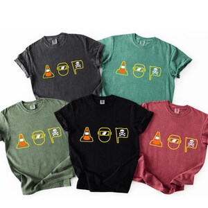 May include: Five short-sleeved t-shirts in charcoal, green, olive, black, and maroon. Each shirt has a yellow graphic with a traffic cone, welding mask, and skull and crossbones, with the letters "AOP".