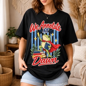 May include: A black short-sleeve t-shirt featuring a graphic of a green frog wearing a red cape and bow tie, holding a yellow highlighter and a blue book. The text reads We Annotate AT Dawn, with a stack of colorful books and blue vertical stripes.