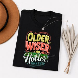 May include: Black t-shirt with the text "Older, Wiser, and Hotter Than Ever" in a colorful design. The text is in yellow, red, and white. A silver necklace with a small pendant is on the shirt.