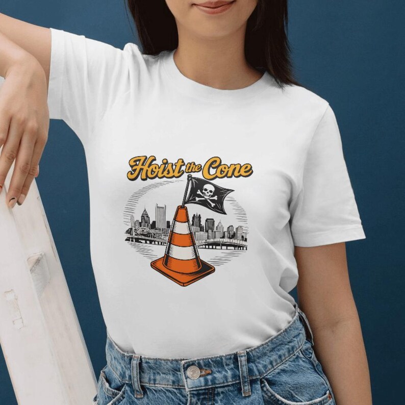 May include: White t-shirt featuring a graphic of a traffic cone with a pirate flag and the text "Hoist the Cone." The design includes a cityscape in the background, with the text in a gold color.