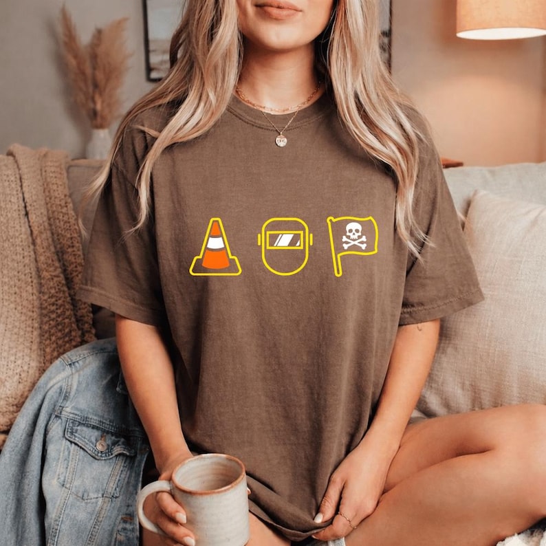 May include: Brown t-shirt with a yellow outline design featuring a traffic cone, a welding mask, and a skull and crossbones flag. The letters "AOP" are implied. The shirt is a relaxed fit.