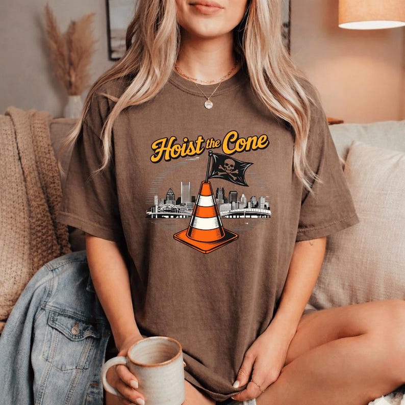 May include: Brown t-shirt with the text "Hoist the Cone" in yellow and a graphic of a traffic cone with a skull and crossbones flag, and a city skyline. The cone is orange and white.