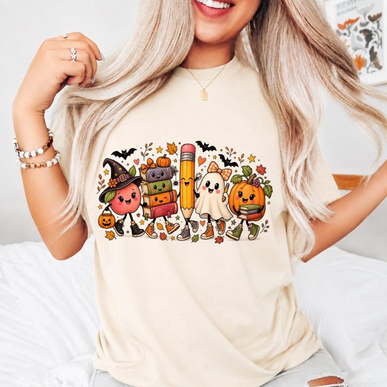 May include: A cream-colored t-shirt featuring a colorful graphic print of a smiling apple wearing a witch hat, a stack of books, a pencil, a ghost, and a pumpkin. The design includes small bats, autumn leaves, and stars scattered around the characters.