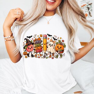 May include: A white t-shirt featuring a colorful graphic of cartoon characters including an apple wearing a witch hat, a stack of books, a pencil, a ghost with a bow, and a pumpkin, all accented with autumn leaves, bats, and stars.