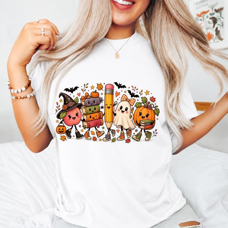 May include: A white t-shirt featuring a colorful graphic of cartoon characters including an apple wearing a witch hat, a stack of books, a pencil, a ghost with a bow, and a pumpkin, all accented with autumn leaves, bats, and stars.