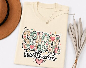 May include: Cream-colored t-shirt with the words "SCHOOL health aide" in a colorful, whimsical design. The design includes hearts, bandages, and a stethoscope. A brown hat and necklace are visible in the background.