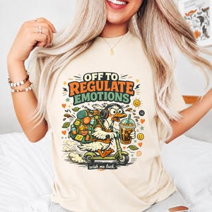 May include: A cream-colored t-shirt featuring a graphic of a white duck wearing headphones, carrying a large backpack, and riding a green scooter while holding an iced coffee. The text reads OFF TO REGULATE EMOTIONS and wish me luck.