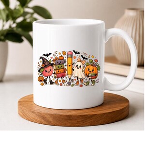 May include: A white ceramic mug featuring a colorful graphic of a smiling apple wearing a witch hat, a stack of books, a pencil, a ghost, and a pumpkin. The characters have arms and legs and are surrounded by small bats and autumn leaves.