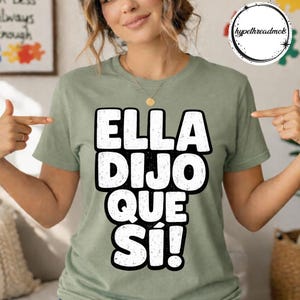 May include: Sage green t-shirt with the text "ELLA DIJO QUE SÍ!" in bold white letters. The shirt has a classic crew neck and short sleeves. A gold necklace is visible.