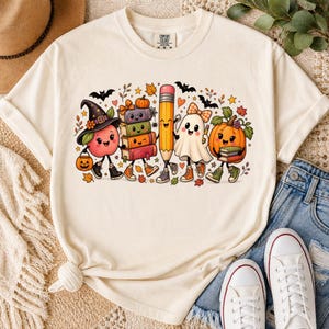 May include: A cream-colored t-shirt featuring a colorful graphic of a smiling apple wearing a witch hat, a stack of books, a pencil, a ghost, and a pumpkin. The design includes small bats, autumn leaves, and hearts scattered around the characters.