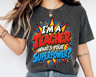May include: A charcoal gray short-sleeve t-shirt featuring a comic book style graphic with a yellow and red starburst background. The text reads "I'M A TEACHER WHAT'S YOUR SUPERPOWER?" in bold white, red, and blue lettering with a dotted pattern.