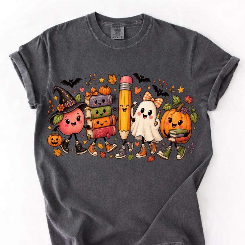 May include: A charcoal gray t-shirt featuring a colorful graphic of anthropomorphic Halloween characters including a witch apple, a stack of books, a pencil, a ghost with a bow, and a pumpkin, all accented with bats, stars, and autumn leaves.