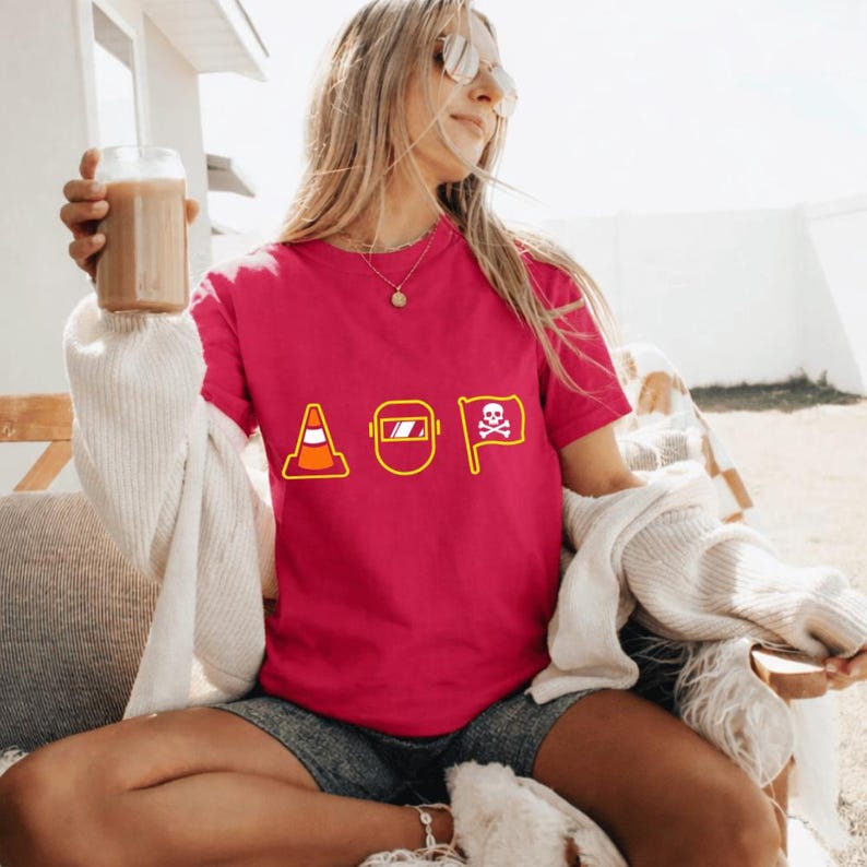 May include: A magenta t-shirt featuring the letters AOP in yellow, each letter represented by a different graphic: a traffic cone, a welding mask, and a flag with a skull and crossbones. The shirt is worn by a person.