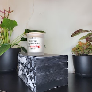 May include: A stack of black books with a white floral design on the side, topped with a candle. The candle has text that reads "Ignoring responsibilities and reading all day." Potted plants and a neon pink light are in the background.