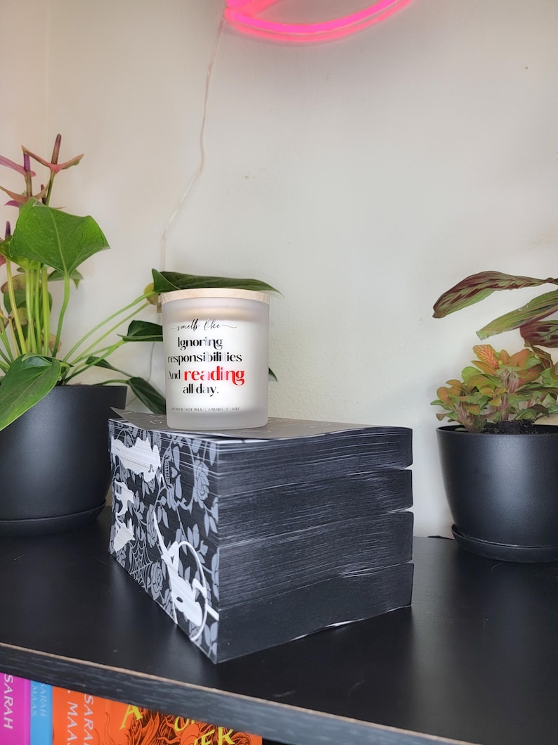 May include: A stack of black books with a white floral design on the side, topped with a candle. The candle has text that reads "Ignoring responsibilities and reading all day." Potted plants and a neon pink light are in the background.