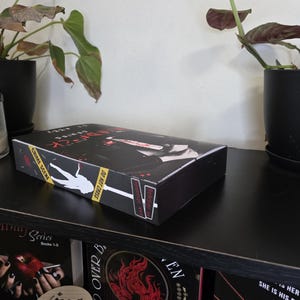 以下が含まれることがあります： 犯罪現場をテーマにした黒い本のボックスセット。シルエットと「CRIME SCENE DO NOT CROSS」の文字が特徴です。ボックスセットには「V」のラベルが付いており、他の本や植物と一緒に黒い棚に置かれています。