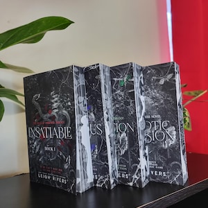 May include: A collection of four books from the "The Darkest Trilogy" series. The book covers are black with white and gray floral and abstract designs. The first book is titled "Insatiable" and the author is Leigh Rivers.