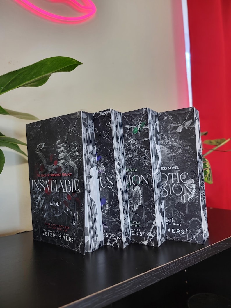 May include: A collection of four books from the "The Darkest Trilogy" series. The book covers are black with white and gray floral and abstract designs. The first book is titled "Insatiable" and the author is Leigh Rivers.