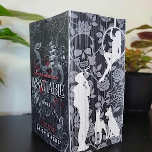 May include: A book titled "Insatiable Book 1" from "The Edge of Darkness Trilogy" by Leigh Rivers. The book has a black and gray cover with a skull, roses, and silhouette figures. The spine features the title and author.