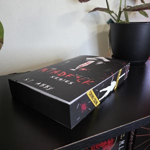 以下が含まれることがあります： S.T. Abbyによる「The Mindf*ck Series」というタイトルの、犯罪現場をテーマにした黒い本のボックスセット。ボックスセットには、人物のシルエットと血しぶきのグラフィックが描かれています。ボックスセットは黒い棚に置かれています。