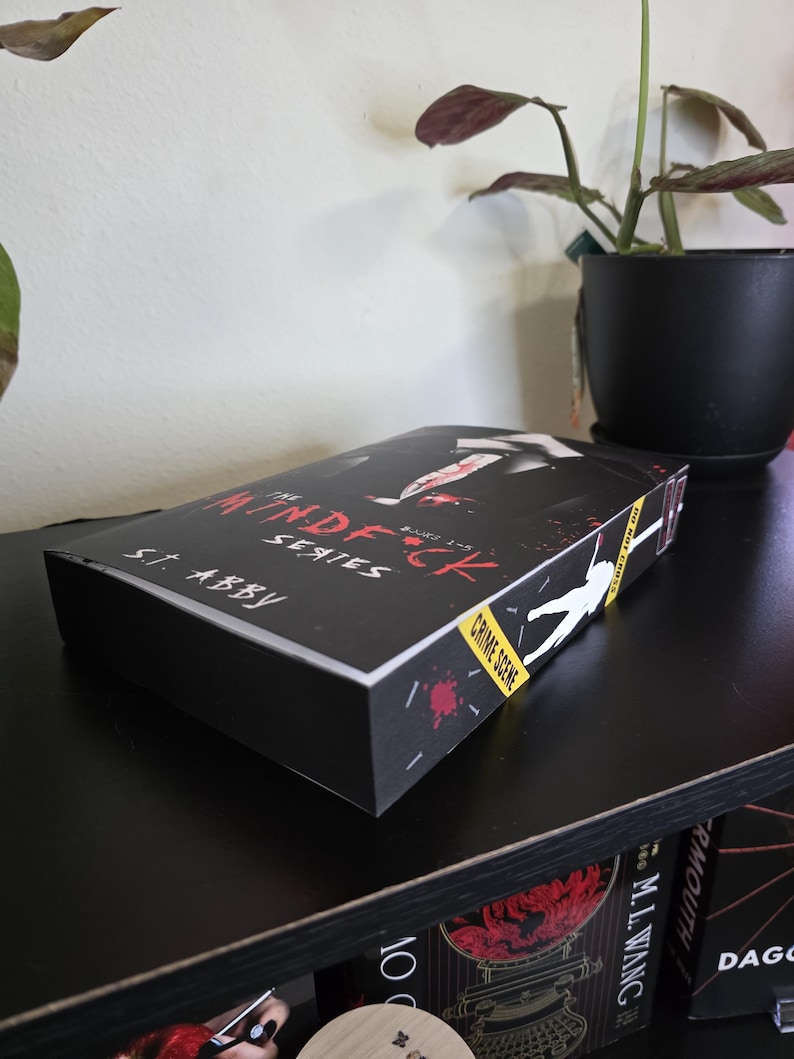 以下が含まれることがあります： S.T. Abbyによる「The Mindf*ck Series」というタイトルの、犯罪現場をテーマにした黒い本のボックスセット。ボックスセットには、人物のシルエットと血しぶきのグラフィックが描かれています。ボックスセットは黒い棚に置かれています。