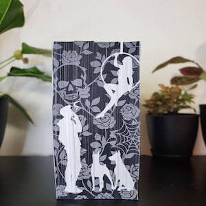 May include: A black and gray decorative book with a skull, roses, a woman in a hoop, a person, dogs, and a spiderweb design. The book is standing upright on a black surface.