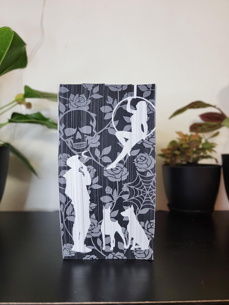 May include: A black and gray decorative book with a skull, roses, a woman in a hoop, a person, dogs, and a spiderweb design. The book is standing upright on a black surface.