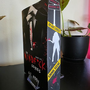 以下が含まれることがあります： S.T. Abbyの「The Mindf*ck Series」というタイトルの本。表紙には血まみれのナイフを持ったスーツ姿の人物が描かれています。背表紙には、シルエットと「Do Not Cross」テープを使った「Crime Scene」のデザインが施されています。