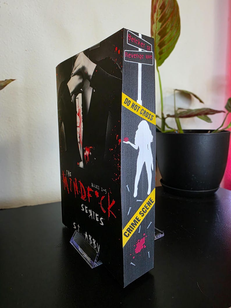 以下が含まれることがあります： S.T. Abbyの「The Mindf*ck Series」というタイトルの本。表紙には血まみれのナイフを持ったスーツ姿の人物が描かれています。背表紙には、シルエットと「Do Not Cross」テープを使った「Crime Scene」のデザインが施されています。