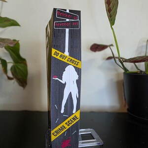 以下が含まれることがあります： 犯罪現場をテーマにした、黒とグレーの装飾ボックス。ボックスには、人物の白いシルエット、"DO NOT CROSS"と"CRIME SCENE"の文字が入った黄色の注意テープ、"Belcher st"と"Revenge ave"と書かれた街路標識が特徴です。