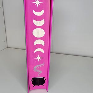 May include: The spine of a pink book with a white star, crescent moons, and a black cauldron with steam coming out of it.