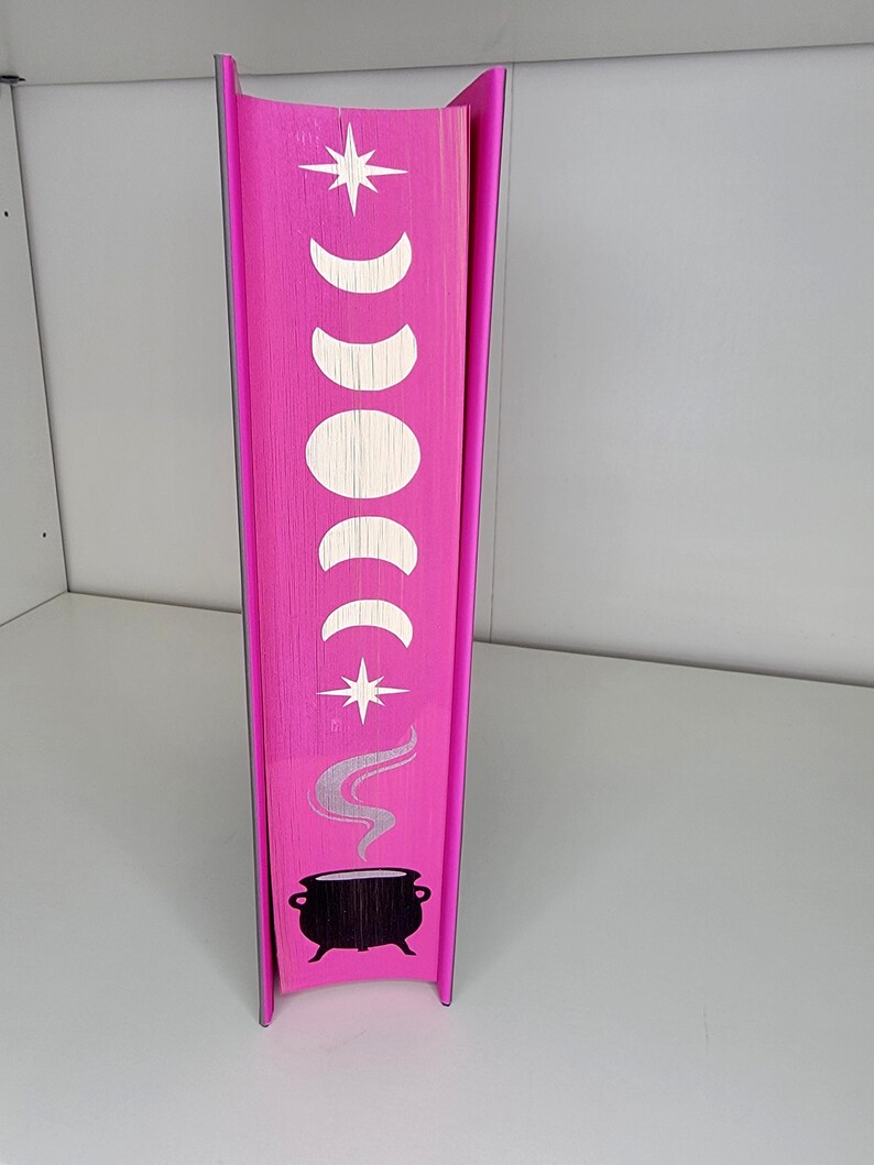 May include: The spine of a pink book with a white star, crescent moons, and a black cauldron with steam coming out of it.