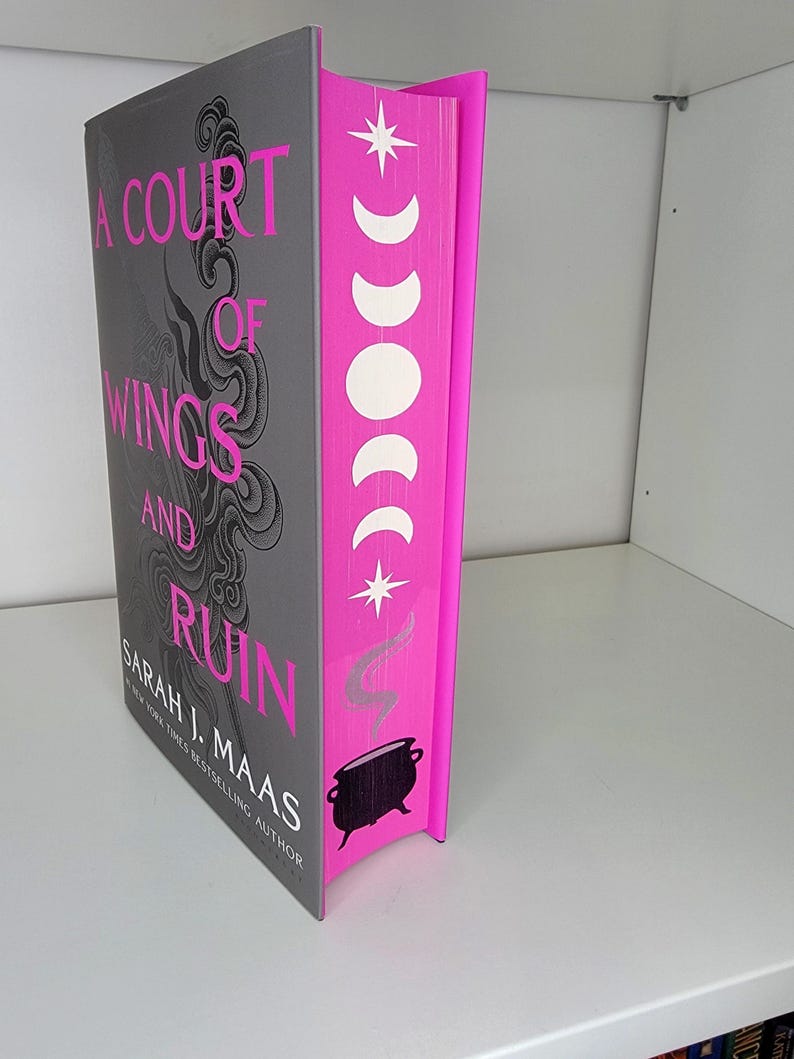 May include: A Court of Wings and Ruin by Sarah J. Maas, a fantasy novel with a pink and gray cover featuring a cauldron and a crescent moon design.