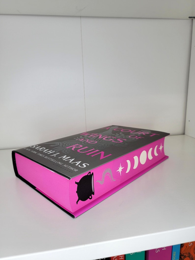 May include: A pink hardcover book with a gray spine and the title "A Court of Wings and Ruin" by Sarah J. Maas. The book cover features a black cauldron with a white star and crescent moons.