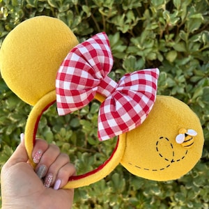 May include: Yellow mouse ear headband with a red and white gingham bow. One ear features a bee applique with a dotted flight path. The headband is yellow with a red interior.