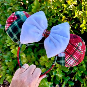 May include: A festive headband with plaid ears in red, green, and white, and a large white bow. The headband is red and green. The ears have a patchwork design. The bow is made of soft, plush material. Perfect for holiday celebrations.