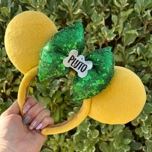 Puede incluir: Diadema de orejas de ratón amarilla con un lazo verde con lentejuelas. El lazo presenta un dije blanco en forma de hueso con el nombre "Pluto" en letras negras. Las orejas y la diadema son amarillas.
