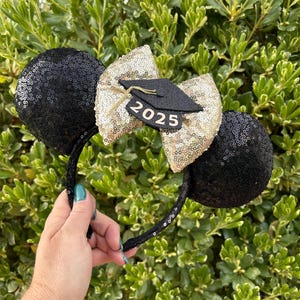 May include: Black sequined mouse ear headband with a gold sequined bow and a small black graduation cap. The bow features the year "2025" in black. The headband is a celebratory accessory.