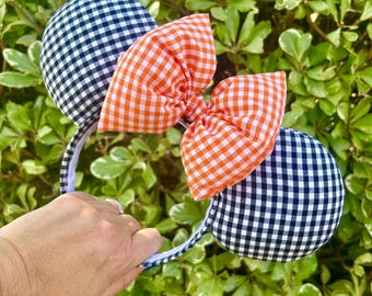 May include: A headband featuring two black and white checkered mouse ears and an orange and white checkered bow. The headband is held in front of a green leafy background.