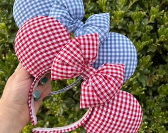 May include: Three headbands with mouse ears and bows. Two headbands have red and white gingham ears and bows. The third headband has blue and white gingham ears and a bow. The headbands are held in front of a green leafy background.