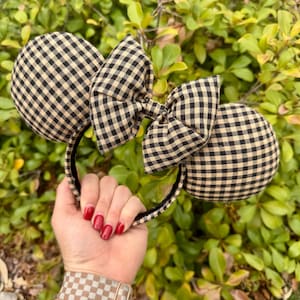 May include: Black and tan gingham pattern mouse ears headband with a matching bow. The ears are round and the bow is centered on the headband. The headband is held in front of a green leafy background.